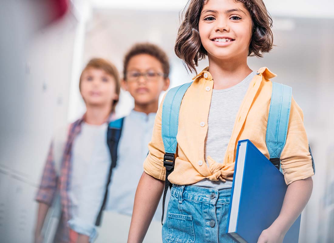 School Insurance - Smiling Young Schoolgirl Walking past Lockers with a Backpack and Carrying Books with Classmates Following behind and Blurred in the Background